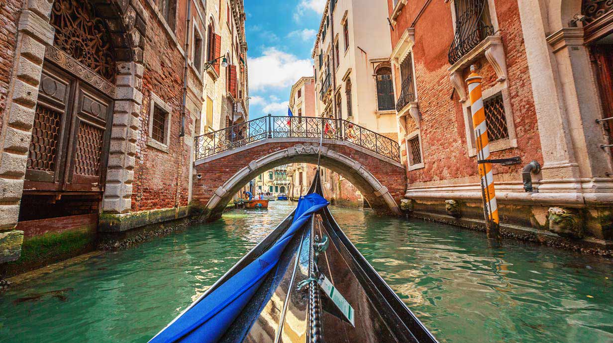 View from a blue gondola with arched stone bridge over canal in background.