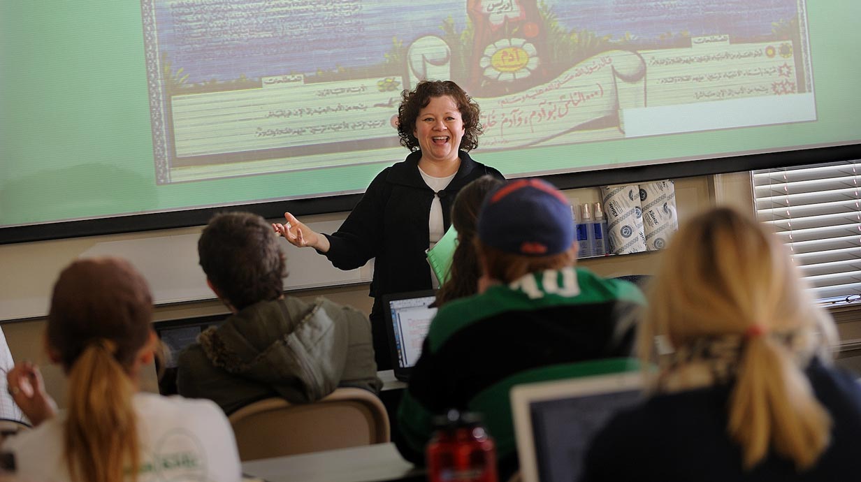 Professor with right arm outstretched lectures to class standing in front of brightly lit screen with illustration and bits of Arabic writing visible. Backs of seated students in foreground.