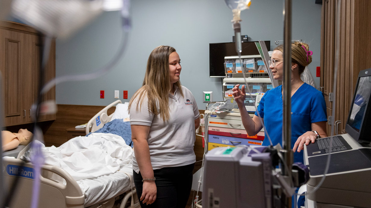 Society and Health student listening to a nurse in hospital room. There is lots of medical equipment in the room and a medical dummy in the bed.