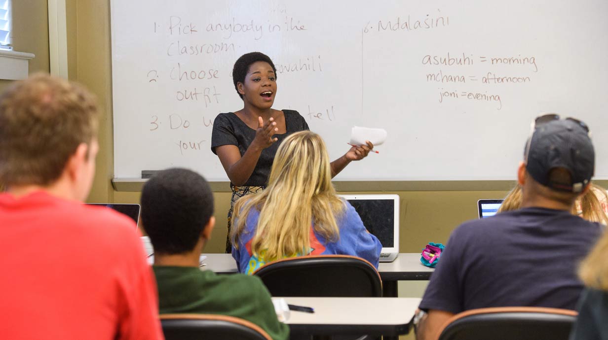 Teacher lecturing to class in front of whiteboard with Swahili written on it.